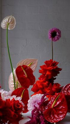 red and white flowers in a vase on a table with brick wall behind them,