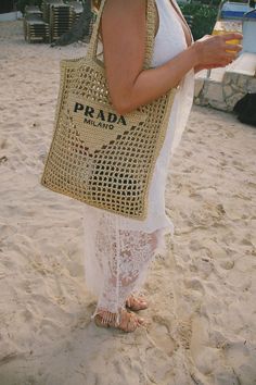 a woman standing on the beach holding a bag and a drink in her hand while wearing sandals