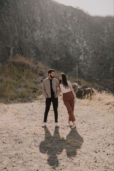 a man and woman standing in the middle of a dirt field next to a mountain