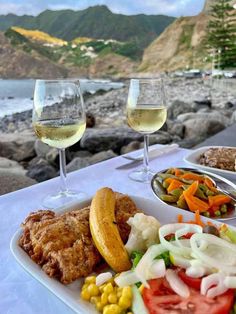 two glasses of white wine are next to a plate of food on a table near the water