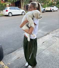 a woman holding a white dog in her arms on the sidewalk next to a street