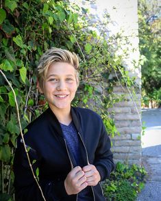 a young boy standing in front of a bush smiling at the camera with his hands on his hips