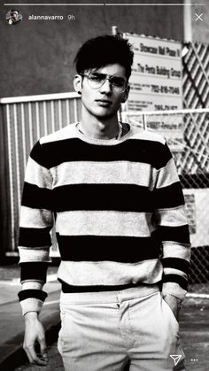 black and white photograph of a young man in striped sweater standing on sidewalk next to fence