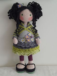 a crocheted doll with black hair wearing a green dress and sandals standing next to a white wall