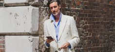 a man in a white suit and blue shirt holding a cell phone next to a brick wall