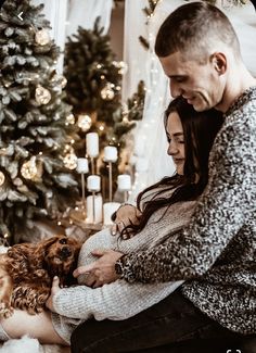 a man and woman sitting on the floor with their dog in front of a christmas tree