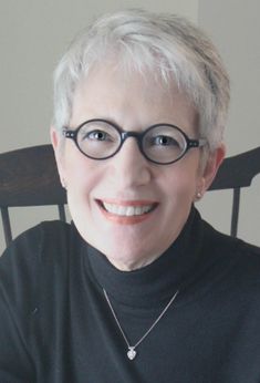 an older woman wearing glasses sitting in a chair with her hands on her hips and smiling at the camera