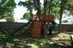 a wooden play structure with a green slide in the back yard next to a tree