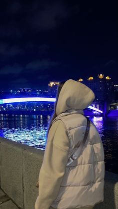 a person wearing a hooded jacket looking at the water and bridge in the distance with lights on