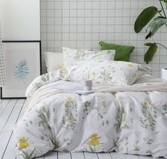 a white bed with yellow flowers and green leaves on the comforter, next to a potted plant