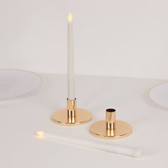 two white candles with gold holders and one candle on a white surface next to a pen