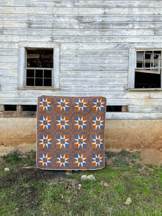 an orange and blue quilt sitting on the ground in front of a white building with two windows