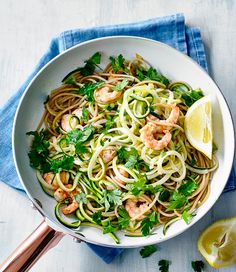 a white bowl filled with pasta and shrimp on top of a blue towel next to a lemon wedge