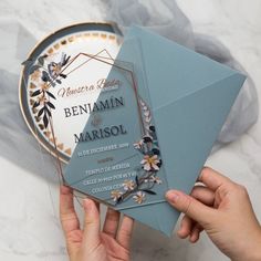 a person holding up a blue and gold wedding card in front of a plate with flowers on it