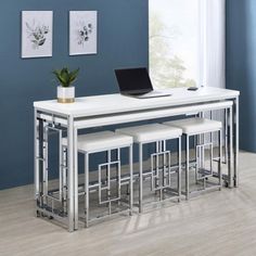 a white dining table with four stools and a laptop on the top, in front of a blue wall