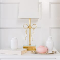 a table with a lamp, vases and a book on top of it in front of a white wall