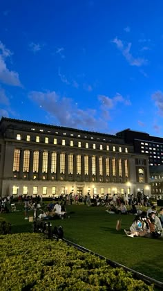 people sitting on the grass in front of a large building at night with windows lit up