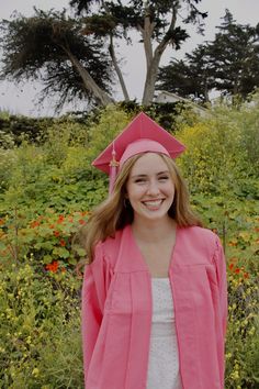 a young woman wearing a pink graduation gown and cap standing in front of some flowers