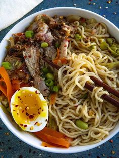 a white bowl filled with noodles, meat and veggies next to an egg