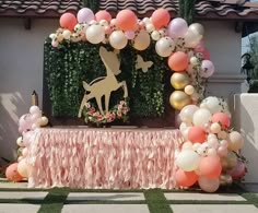 a table topped with balloons and a deer statue next to a wall covered in greenery