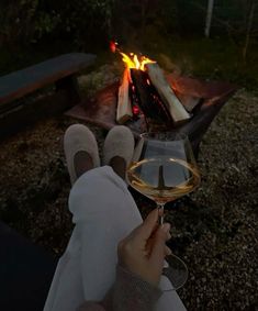 a person holding a glass of wine in front of a campfire with their feet up