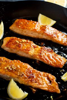 two salmon fillets cooking in a skillet with lemon wedges on the side