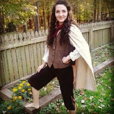 a woman standing in front of a fence with a cape over her shoulders and boots on