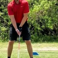 a man in red shirt and black shorts holding golf club on grass with trees in background