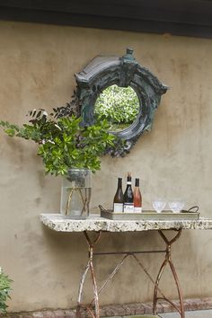 a table with bottles and glasses on it in front of a wall mounted mirror, next to a potted plant