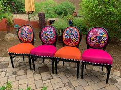 four colorful chairs sitting on top of a brick floor next to trees and bushes in the background