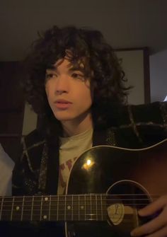 a young man with curly hair playing an acoustic guitar in his bedroom at night time