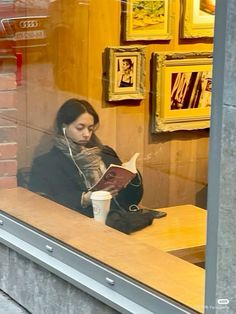 a woman sitting at a table reading a book and drinking coffee in front of a window