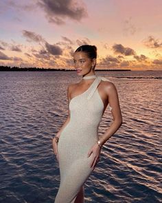 a woman in a white dress standing on the edge of a body of water at sunset