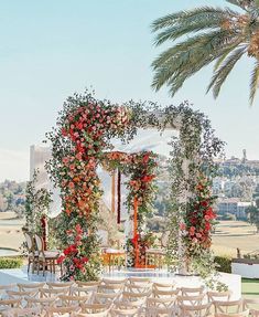 an outdoor wedding setup with white chairs and red flowers on the arch, surrounded by palm trees