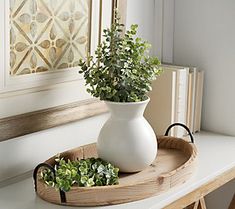 two white vases sitting on top of a wooden tray with plants in them next to a window