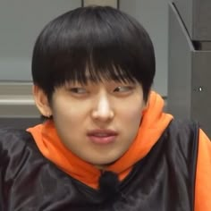 a young man with black hair and an orange hoodie looks at the camera while sitting down