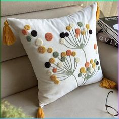 a white pillow with flowers and tassels sitting on a couch next to a pair of glasses