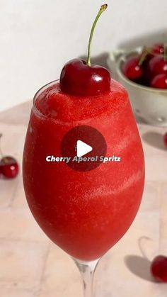 a red drink sitting on top of a table with cherries in the bowl behind it