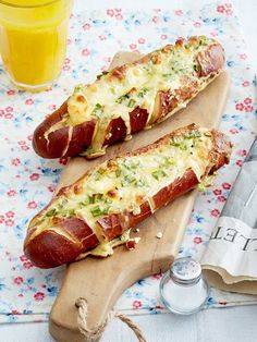 two hot dogs with cheese and toppings are on a wooden board next to a glass of orange juice