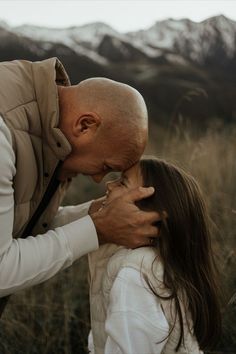 an older man kissing a young woman on the forehead in a field with mountains in the background