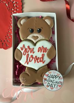 a teddy bear holding a heart shaped cookie in a box with the words you are loved written on it