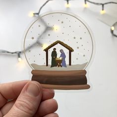 a hand holding up a snow globe with a nativity scene on it and lights in the background