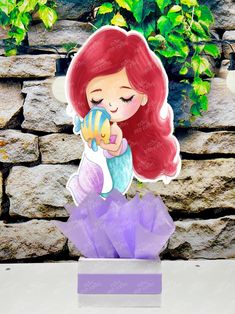 the little mermaid is holding a fish in her hand while sitting on top of a purple box