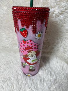 a pink cup with strawberries on it is sitting on a white blanket and has a straw