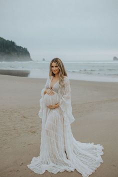 a pregnant woman standing on the beach wearing a white dress and holding her belly up