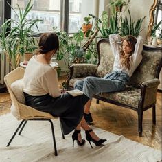 two women sitting on chairs in a room with plants and potted plants behind them