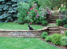 a black cat sitting on top of a stone wall in a garden next to flowers