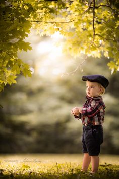 a little boy standing under a tree in the grass wearing a hat and plaid shirt