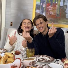 two people sitting at a table with food and drinks in front of them, making the peace sign