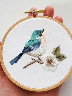 a hand holding a small embroidered bird on a branch with flowers in front of it